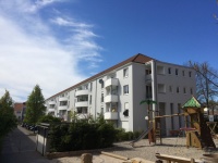 Wohnhäuser mit Satteldach, graue Fassade, weiße Balkonbrüstungen. Parkplätze mit Autos, rechts Spielplatz. 