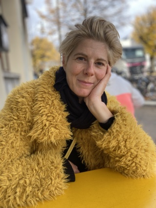 Portraitfoto von der Regisseurin Birgit Möller, draußen, den Kopf mit der Hand abgestützt, in gelber Teddyjacke