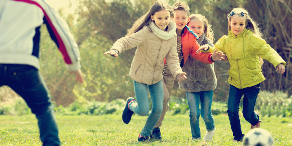 Vier Kinder rennen auf einen Fußball zu. Ein Kind steht mit dem Rücken zur Kamera.