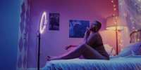 Frau sitzt mit Unterwäsche auf einem Bett vor einem Licht und ihrem Handy