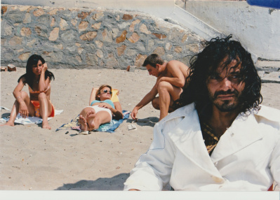 Junger Mann im weißen Mantel am Strand. Hinter ihm sitzt eine Frau auf ihrem Handtuch, eine weitere liegt auf ihrem und neben ihr kniet ein anderer junger Mann