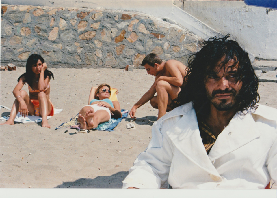 Junger Mann im weißen Mantel am Strand. Hinter ihm sitzt eine Frau auf ihrem Handtuch, eine weitere liegt auf ihrem und neben ihr kniet ein anderer junger Mann