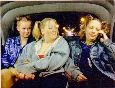 3 Jugendliche sitzen hinten im Auto. Eine telefoniert und schaut genervt, die anderen beiden gucken sie lachend an