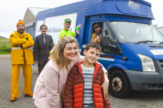 Eine Frau kniet neben einem Jungen, hat ihren Arm um ihn gelegt. Im Hintergrund ein kleiner Truck, aus dem ein Mann rausschaut. Daneben 3 weitere Männer in Arbeitskleidung, Anzug und Sportkleidung