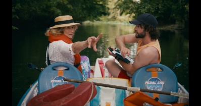 Eine ältere Dame mit Sonnenhut und ein junger Mann mit Klemmbrett und Stoppuhr sitzen nebeneinander in einem Tretboot. Der Mann schaut die Dame an und sie zeigt und schaut hinter die beiden. Hintergrund ist eine Flusslandschaft