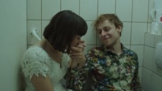 Ein junger Mann und eine junge Frau sitzen im Badezimmer. Die Frau küsst seine Hand, er lächelt sie an