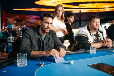 2 Männer am Pokertisch in einem Casino. Beide schauen geradeaus, der eine hebt seine Karten an
