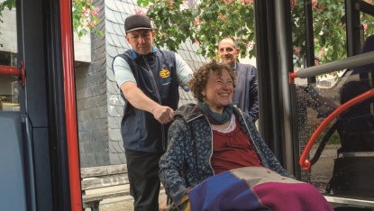 Auf dem Foto sieht man einen mobisaar-Lotsen, der eine Kundin im Rollstuhl in den Bus schiebt.  Im Hintergrund ist noch ein mobisaar-Lotse