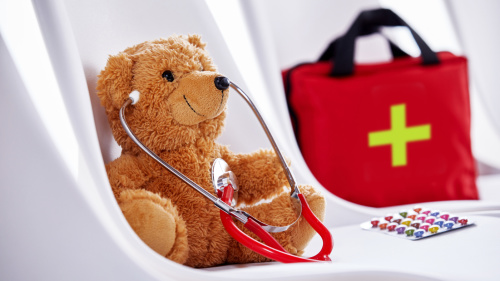 Das Bild zeigt einen braunen Teddybären, der auf einem weißen Stuhl sitzt und ein rotes Stethoskop um den Hals trägt. Im Hintergrund steht eine rote Erste-Hilfe-Tasche mit einem grünen Kreuz. Auf dem Stuhl liegt neben dem Bären eine Packung bunter Pillen.