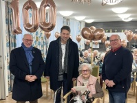 Drei Männer neben einer Seniorin im Rollstuhl mit goldenen Ballons und der Aufschrift "Happy Birthday", vor einem Raum mit sitzenden Menschen