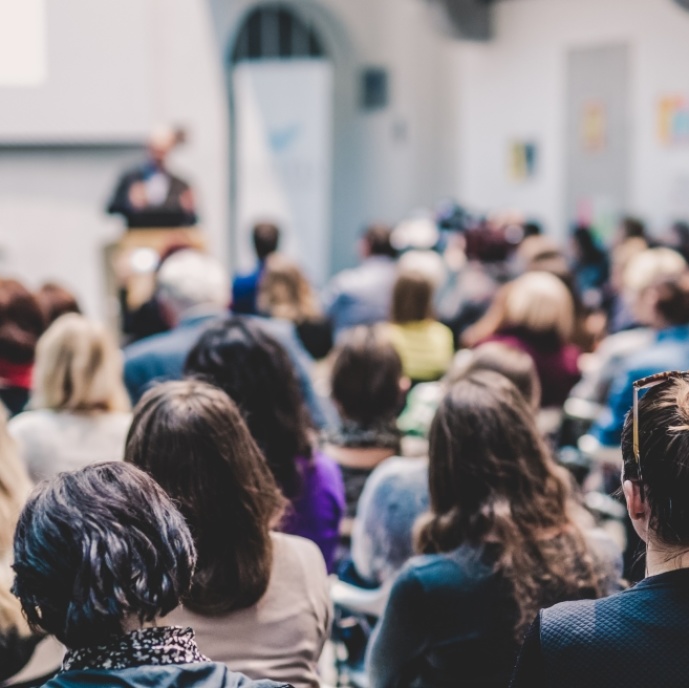 Publikum sitzt in einer Vortragshalle, im Hintergrund steht ein Referent an einem Pult