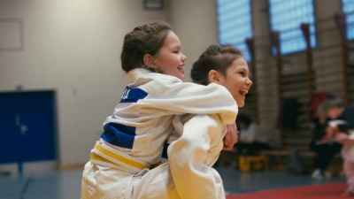 Zwei Mädchen im Sportunterricht. Ein Mädchen sitzt auf dem Rücken der anderen. Beide sind am lachen.