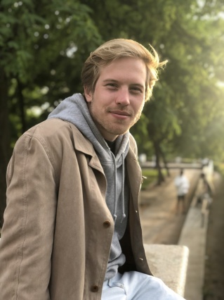 Junger Mann mit kurzen blonden Haaren. Er sitzt auf einer Mauer und schaut in die Kamera.