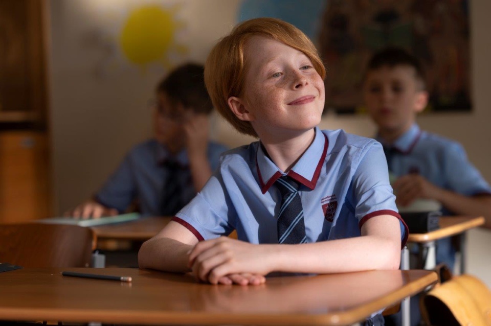 Ein Junge in Schuluniform sitzt in einem Klassenzimmer, lächelt und schaut nach links.