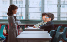 Drei Kinder in einem Klassenzimmer. Zwei Jungen sitzen an ihren Tischen und ein Mädchen steht mit verschränkten Armen vor ihnen.