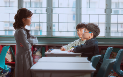 Drei Kinder in einem Klassenzimmer. Zwei Jungen sitzen an ihren Tischen und ein Mädchen steht mit verschränkten Armen vor ihnen.