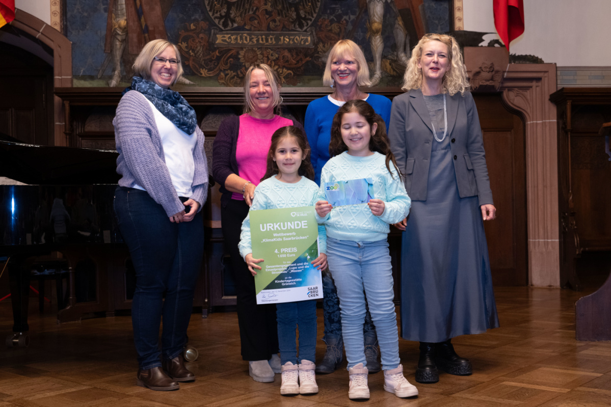 Preisübergabe des vierten Platzes an die Kita Grüneich. Zu sehen sind zwei Kinder welche die Urkunde halten und 2 Betreuerinnen der Kita, sowie zwei Jurymitglieder.