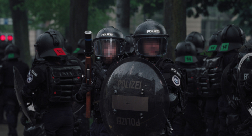 Zwei Polizisten in Uniform und schauen in die Kamera. Einer hält ein Schutzschild vor sich. Beide Gesichter sind verpixelt. Hinter ihnen sieht man noch weitere Polizist:innen