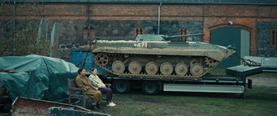 Zwei Frauen sitzen auf einer Bank. Neben ihnen steht ein Panzer auf einem Wagen.