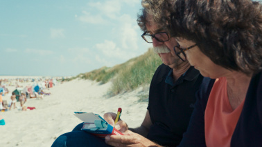 Ein Mann und eine Frau sitzen an Dünen am Strand. Er notiert etwas und sie schaut dabei zu.