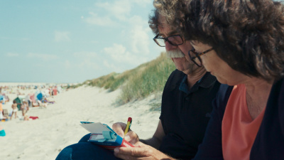 Ein Mann und eine Frau sitzen an Dünen am Strand. Er notiert etwas und sie schaut dabei zu.