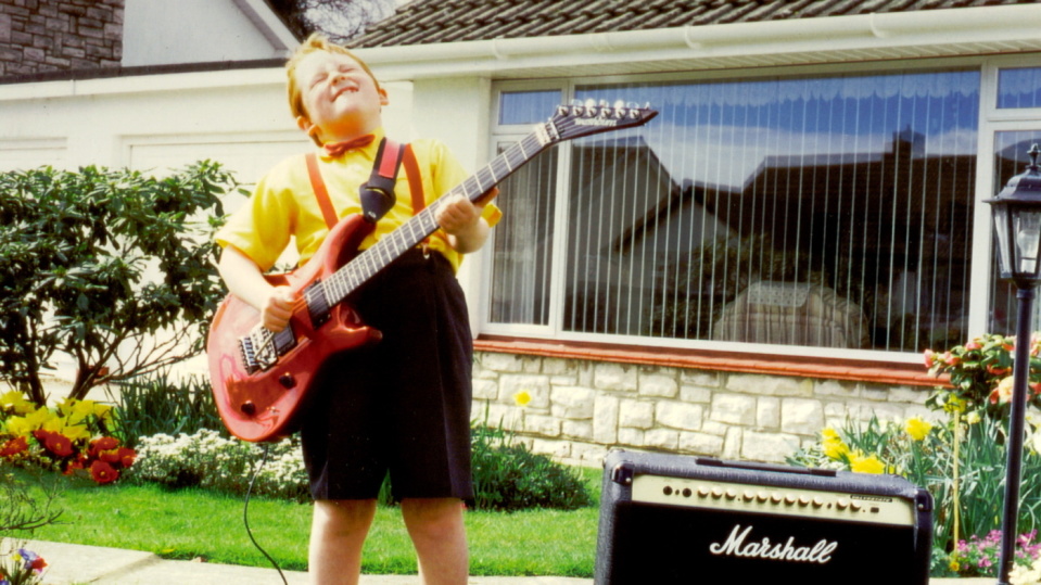 Ein Junge in Hemd und Hosenträgern spielt mit geschlossenen Augen eine E-Gitarre im Vorgarten vor einem Haus.