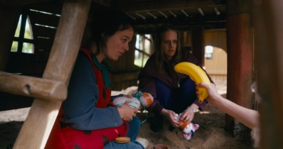 Zwei Frauen sitzen unter einem Bauwerk aus Holz. Auf eine der Frauen wird mit einer Bananen-Aufbewahrungsdose gezeigt.