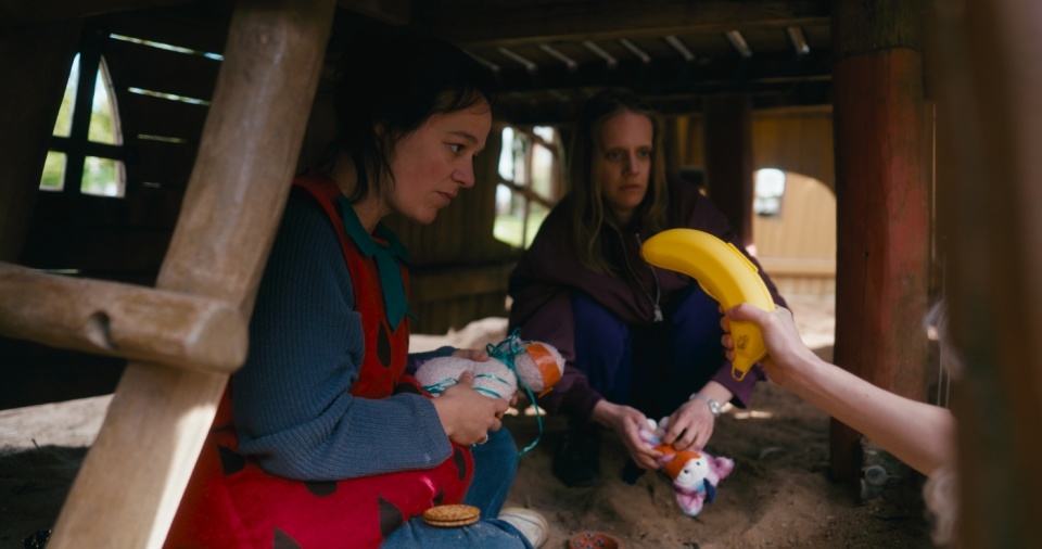 Zwei Frauen sitzen unter einem Bauwerk aus Holz. Auf eine der Frauen wird mit einer Bananen-Aufbewahrungsdose gezeigt.