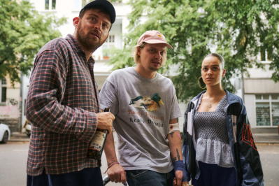 Drei junge Menschen stehen vor einer urbanen Kulisse, die mit ernstem Blick auf etwas hinter der Kamera schauen.