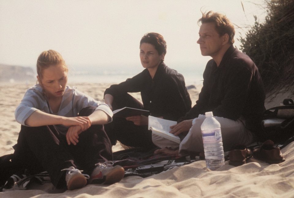 Ein Mann und zwei Frauen sitzen an einem Strand. Eine Frau schaut vor sich auf den Boden, die anderen beiden schauen in die Ferne. Die Stimmung scheint angespannt. 