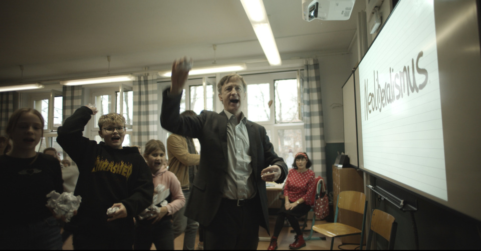 In einem Klassenzimmer stehen ein Lehrer im Anzug und mehrere Kinder, die Papierbälle werfen und jubeln. Im Hintergrund ist eine beschriebene Tafel mit dem Wort „Neoliberalismus“ zu sehen. Die Szene wirkt lebendig und chaotisch.