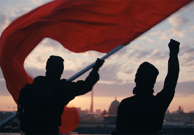 Zwei dunkel gekleidete Personen stehen vor einem Sonnenuntergang, eine hält eine große rote Fahne, die im Wind weht, während die andere die Faust in die Luft streckt. Im Hintergrund ist die Silhouette einer Stadt mit markanten Gebäuden und einem Fernsehtu