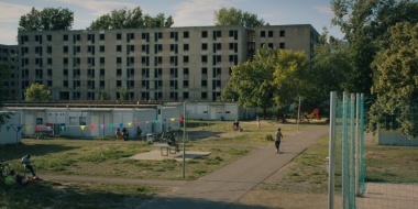 Foto eines Geflüchtetenlagers, bestehend aus Wohncontainern, Spiel- und Sportplätzen, vor verlassenen Hochhäusern.