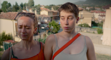Zwei Frauen, im Hintergrund eine italienische Stadt. Die eine kneift ihre Augen zusammen, während die andere sie fragend anschaut.
