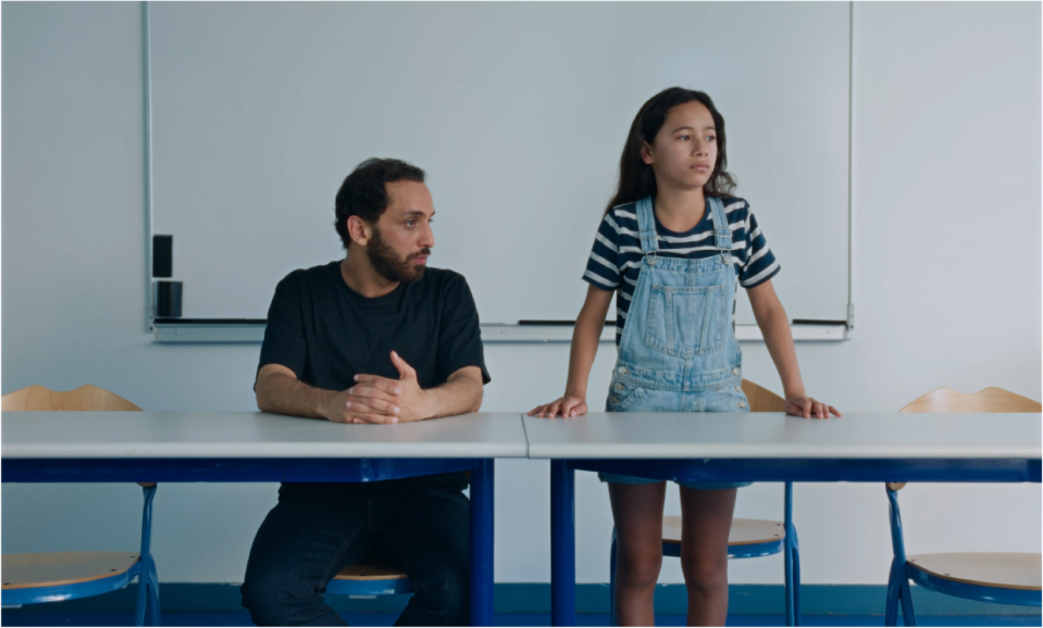 Ein Mann und ein Mädchen nebeneinander im Klassenzimmer. Der Mann sitzt, das Mädchen steht und lehnt auf dem Tisch. Beide schauen ernst in dieselbe Richtung.