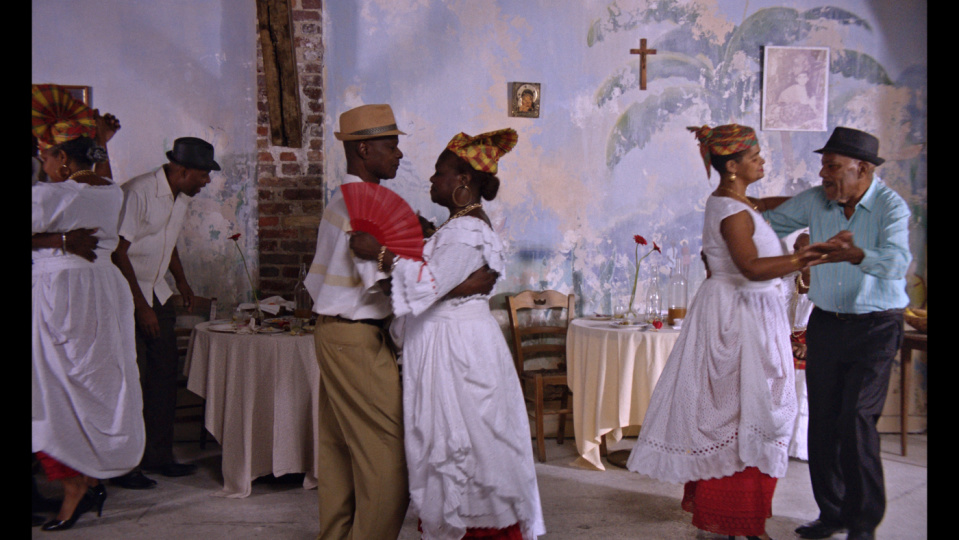 In einem lebhaften Raum tanzen mehrere Paare in traditioneller Kleidung. Der Hintergrund ist von gedeckten Tischen, einer verzierten Wand und dezenten Dekorationen geprägt. Die Szene strahlt Lebensfreude, Tradition und Gemeinschaft aus.