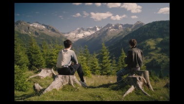 Zwei junge Erwachsene sitzen auf abgesägten Baumstämmen auf einer Berglandschaft und blicken in die Ferne.