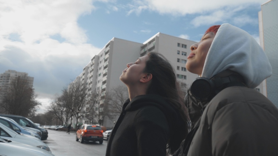 Zwei Personen in einem urbanen Stadtgebiet heben die Köpfe mit geschlossenen Augen gen Himmel.