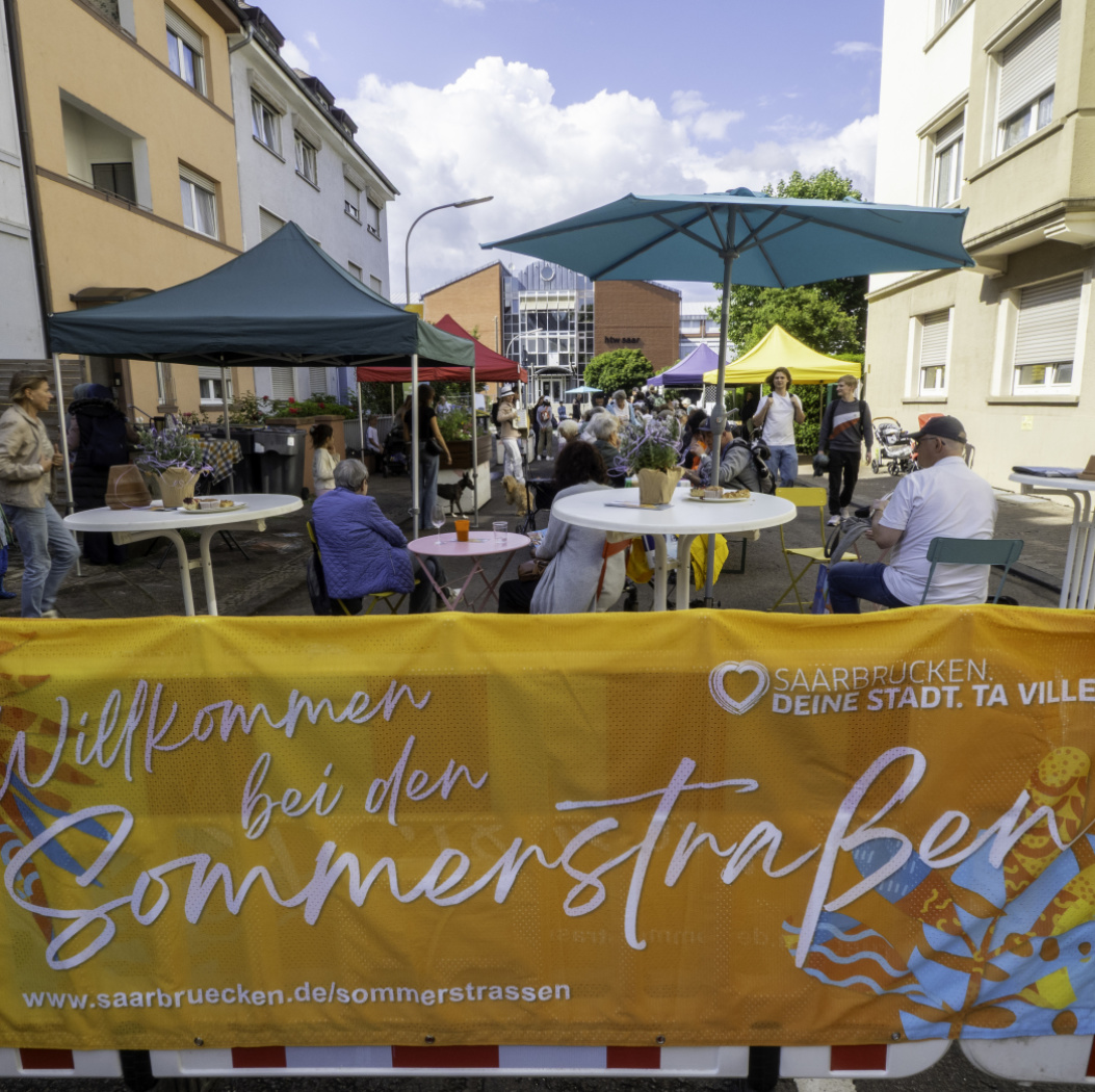 Vorne steht eine Absperrung über die ein Banner gespannt ist. Dahinter ist eine Straße mit vielen Passanten und bunten Schirmen zu sehen.