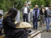 Eine Frau sitzt auf der Bank und spielt auf einer Klangschale. Im Hintergrund stehen Menschen und unterhalten sich