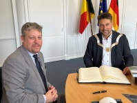 Der belgische Botschafter, S.E. Piet Heirbaut, hat sich bei seinem Besuch im Rathaus am 17. Juli in das Golde Buch der Stadt eingetragen. Zwei Männer sitzen an einem Tisch vor einem großen Buch. Im Hintergrund sind die belgische, die europäische und die deutsche Fahne zu sehen.