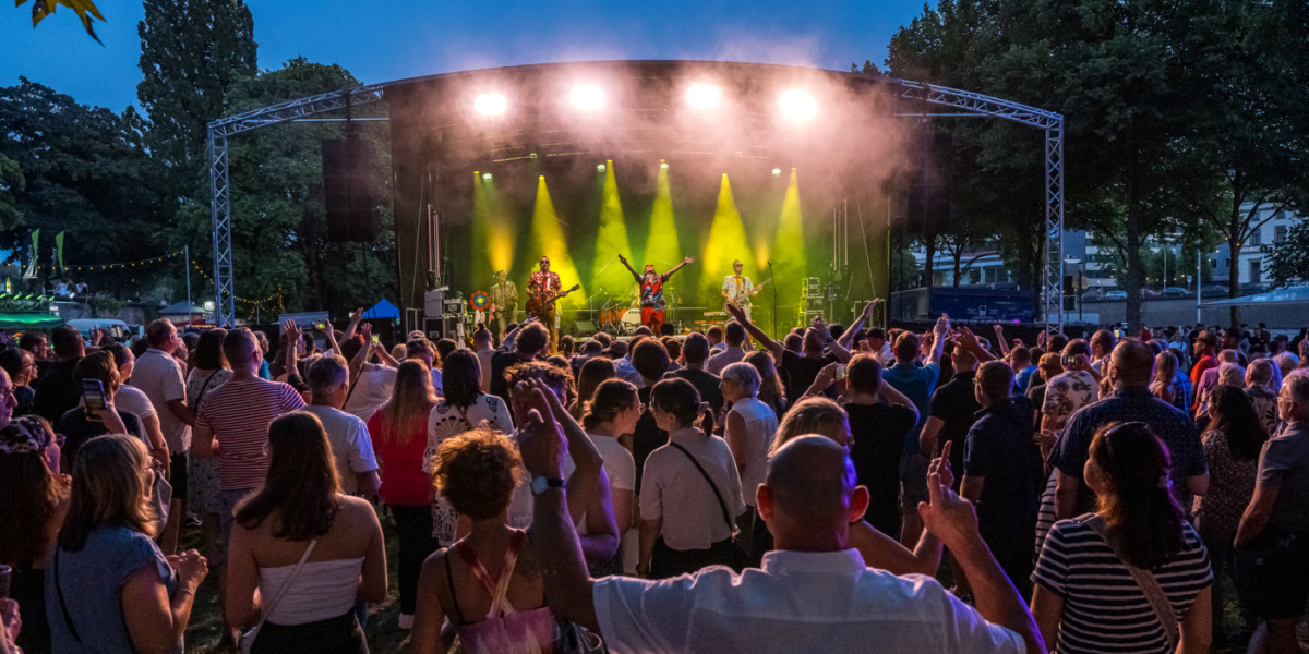 Auf einer gelb erleuchteten Bühne im Hintergrund spielt eine Band, davor steht Publikum mit teilweise tanzenden Menschen.