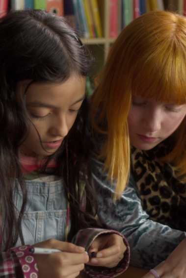 IM KREIS Zwei Mädchen sitzen gemeinsam an einem Tisch und betrachten ein Heft, das vor ihnen liegt.