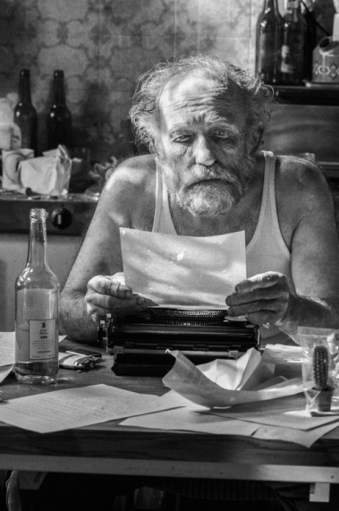 Ein älterer Mann sitzt in einer unordentlichen Küche an einer Schreibmaschine und liest ein Blatt Papier.