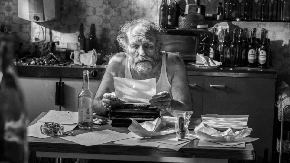 Ein älterer Mann sitzt in einer unordentlichen Küche an einer Schreibmaschine und liest ein Blatt Papier.