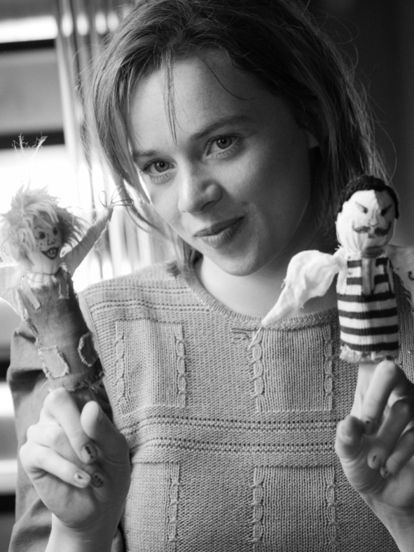 Eine junge Frau steht vor einem Treppenaufgang und hält in jeder Hand eine Fingerpuppe.