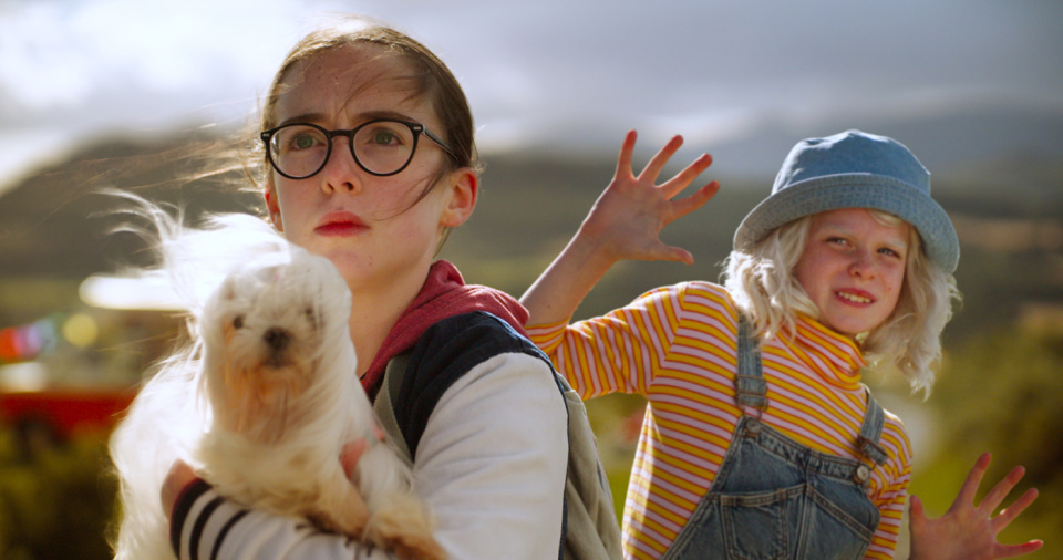 Zwei Kinder stehen draußen; eines hält einen kleinen weißen Hund, das andere macht eine verspielte Handbewegung. Zwei Kinder stehen draußen; eines hält einen kleinen weißen Hund, das andere macht eine verspielte Handbewegung.