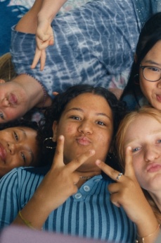 Echt Friends Eine Gruppe jugendlicher Mädchen liegen nebeneinander auf einer Decke, zeigen jeweils mit den Händen ein Peace-Zeichen, umgeben von Kissen und einem Smartphone.