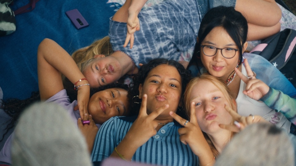 Eine Gruppe jugendlicher Mädchen liegen nebeneinander auf einer Decke, zeigen jeweils mit den Händen ein Peace-Zeichen, umgeben von Kissen und einem Smartphone.