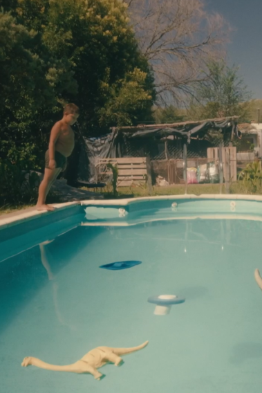 EL MUNDO AL REVÉS Ein Junge steht am Beckenrand eines Swimmingpools in einem Garten. Im Pool schwimmen zwei Spielzeug-Dinosaurier.